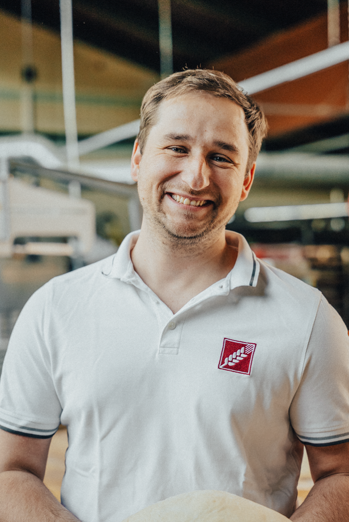 Portrait von Frank Lehrbach in der Backstube. Er trägt Arbeitskleidung und lächelt in die Kamera.