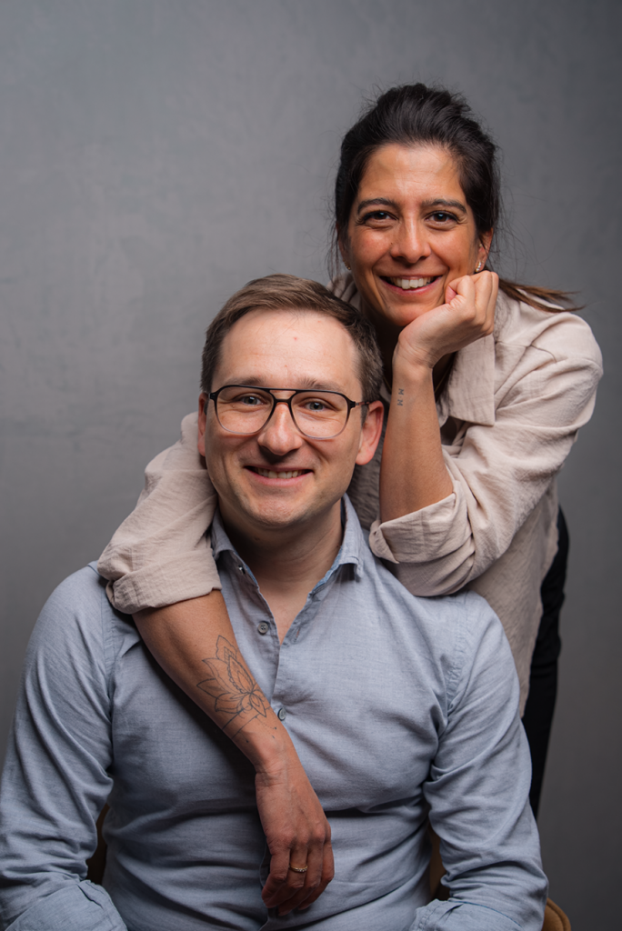 Carina und Frank Lehrbach lächeln in die Kamera. Carina steht umarmend hinter dem sitzenden Frank.