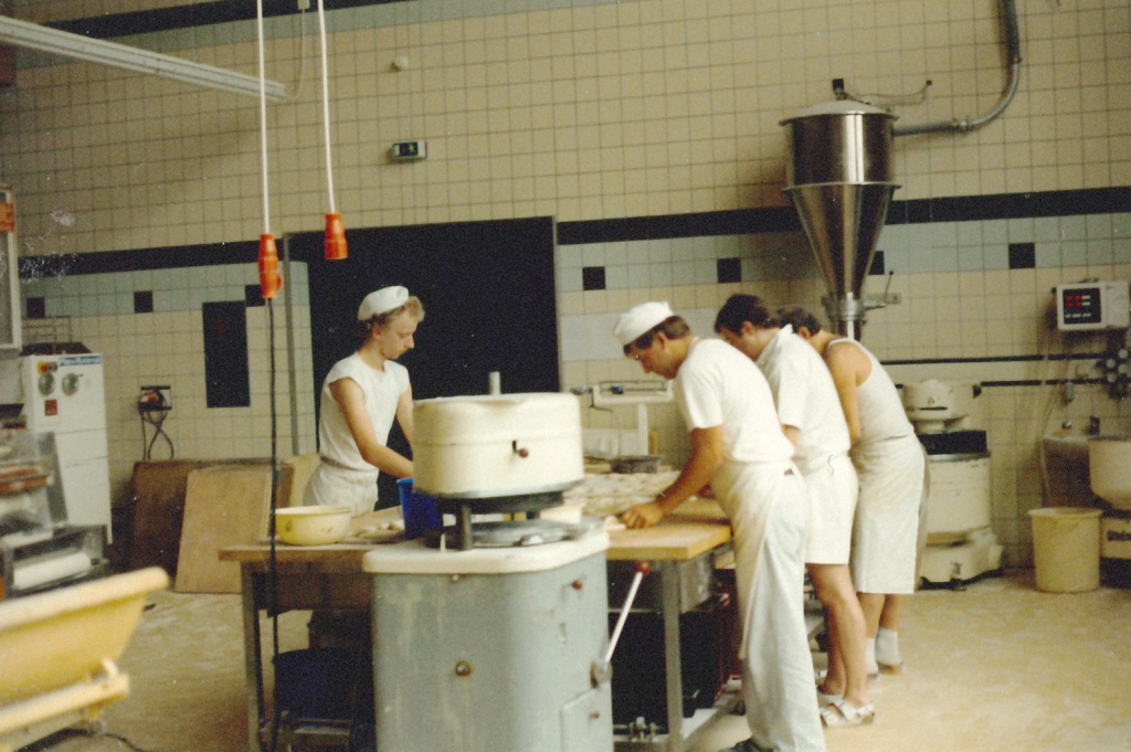 Foto aus der Backstube 1988. 4 Männer stehen in Arbeitskleidung vor einem Tisch und kneten und formen Teig.